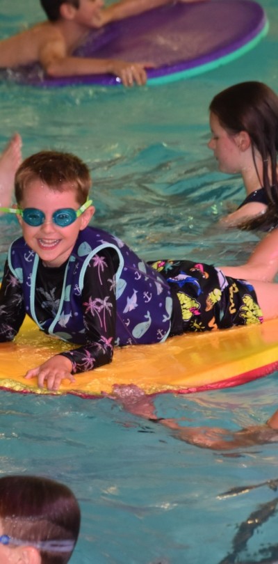 Boy on mat in swimming pool surrounded by swimmers